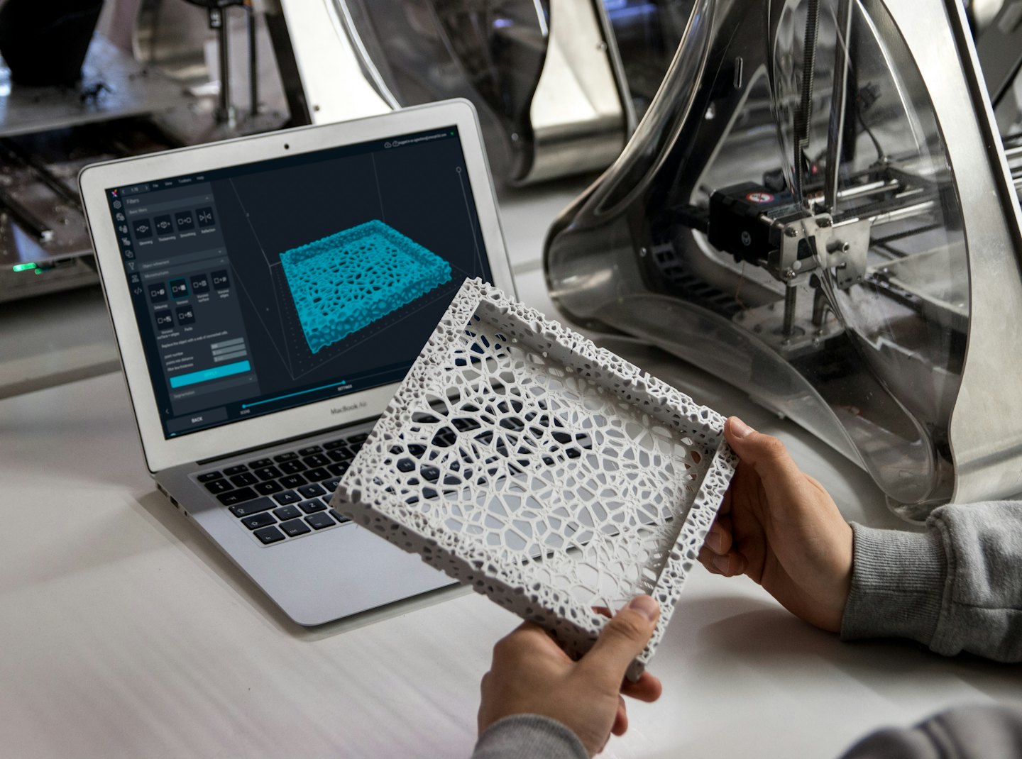 Manos sostienen un objeto impreso en 3D junto a una laptop que muestra un modelo 3D.